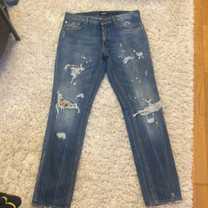 Blå slitna jeans från Just Cavalli - Säljer ett par blå jeans från Just Cavalli med flera slitningar och trasiga detaljer framtill. Jeansen har klassisk femficksdesign, raka ben och normal passform. Perfekta för dig som gillar en edgy och avslappnad stil.