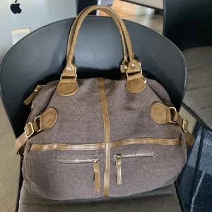 Brun handväska med detaljer i skinn - Säljer en brun handväska med snygga ljusbruna skinn-detaljer och flera dragkedjor framtill. Väskan har två handtag och justerbara spännen på sidorna. Perfekt storlek för att få plats med allt du behöver.