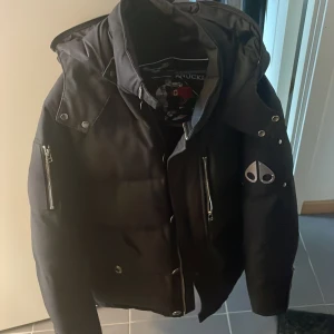 Svart pufferjacka från Moose Knuckles - Säljer en svart pufferjacka från Moose Knuckles med huva och silverfärgade dragkedjor. Jackan har logotyp på ärmen och flera fickor med dragkedja. Perfekt för kalla dagar. Dragkedjan är sönder och ett bränn märke men den är äkta