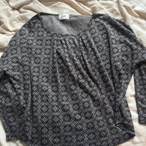 Mönstrad långärmad blus från Isay - Svart och grå klänning från Isay med geometriskt mönster över hela plagget. Långärmad modell med rund halsringning och svarta paneler längs ärmarna. Mjuk och bekväm, perfekt för dig som gillar en unik stil.