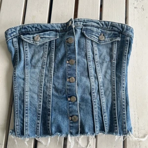 Jeans korsettopp från Zara - Säljer en jeans korsettopp i storlek S från Zara i blå denim. Toppen har knappar framtill, två fejkade bröstfickor och en fransig nederkant. Baksidan har smock för extra stretch och passform. (Toppen är oanvänd)