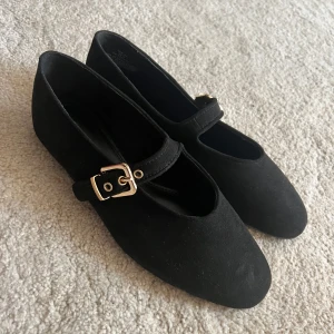 Svarta ballerinaskor med spänne H&M - Stilrena svarta ballerinaskor från H&M med semi-rund tå och snyggt guldfärgat spänne över foten. Skorna är i mockaimitation och har platt sula.