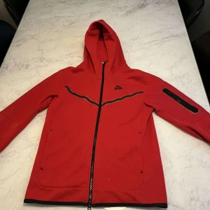 Nike tech set - Helt ny,  Röd hoodie från Nike med hel dragkedja, huva och svarta detaljer. Jackan har svarta Nike-loggan på bröstet, svarta linjer över bröstet och en ficka med dragkedja på ärmen. Perfekt för dig som gillar sportig och clean stil.