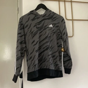 Grå hoodie med zebramönster från Adidas - Snygg grå hoodie från Adidas med svart zebramönster och klassiska vita ränder längs ärmarna. Tröjan har huva, långa ärmar och svart mudd vid ärmslut och nederkant. Perfekt för dig som gillar sportig och trendig stil.