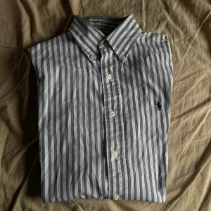 Randig skjorta från Polo Ralph Lauren - Snygg skjorta från Polo Ralph Lauren i vitt med svarta ränder. Klassisk krage, knäppning framtill och broderad logga på bröstet. Tillverkad i 100% bomull för en skön känsla och stilren look.