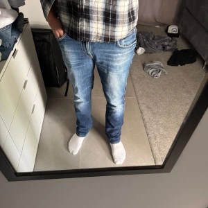 Nudie Jeans  - Säljer ett par Nudie Jeans Slim Jim i tvätten Core Blue. De är blå med slitningar och har klassiska Nudie-detaljer på bakfickorna. Modellen är slim fit med rak passform och tillverkad i 98% bomull och 2% elastan för lite stretch. Storlek 33/32