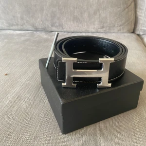 Svart H bälte - Snyggt svart skärp med ikoniskt H-spänne i silverfärgad metall. Bältet är gjort i äkta skinn med vit kontrastsöm längs kanterna. Perfekt accessoar för att lyfta din outfit med en lyxig touch.