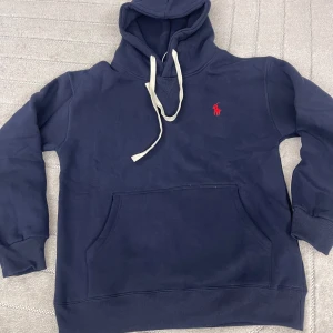 Mörkblå hoodie  - Säljer en mörkblå hoodie  med klassisk röd logg. Tröjan har huva med vita snören, känguruficka och ribbade muddar. Tillverkad i mjuk bomullsmix som är perfekt för kyliga dagar.