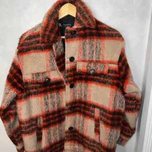 ZARA jacka i ullmix - Overshirt från ZARA i mjuk ullmix med rött, beige och brunt rutmönster. Jackan har stora knappar framtill, bröstfickor med lock och klassisk krage. Perfekt för lager-på-lager och ger en cool vibe till din outfit.