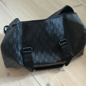 Svart weekendbag med mönster - Stor svart weekendbag med diskret mönster i material som känns som syntet. Väskan har två kraftiga handtag, dragkedja upptill och ett rymligt huvudfack med innerficka. Perfekt för resor eller gymmet när du vill ha plats för allt.