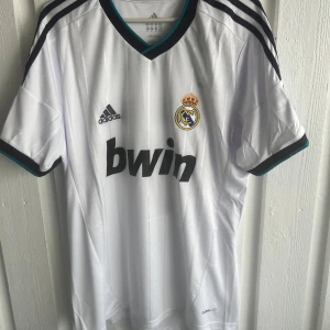 Real Madrid Ronaldo 7 Adidas tröja - Säljer en klassisk Real Madrid fotbollströja med Ronaldo 7 på ryggen. Tröjan är vit med svarta och mörkblå detaljer, Adidas-logga och klubbmärke på bröstet samt Champions League-märke på ärmen. Tillverkad i lätt och ventilerande material (Climacool).