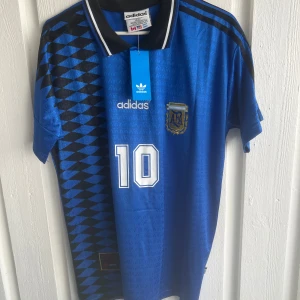 Argentina Maradona 10 blå Adidas tröja - Blå Argentina fotbollströja från Adidas med Maradona och nummer 10 på ryggen. Tröjan har svartvitt krage, svarta ränder på axlarna och ett coolt geometriskt mönster på ena sidan. Broderad landslagssköld och Adidas-logga framtill. Tillverkad i lätt polyester.