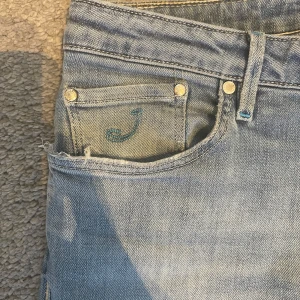 Ljusblå jeans från Jacob Cohën - Snygga ljusblå jeans från Jacob Cohën med klassisk femficksdesign, ljusblå broderad J på myntfickan och patch i mocka baktill. Jeansen har silverfärgade nitar och knapp, samt blå detaljer vid knapphål och bälteshällor. Perfekta för en avslappnad och trendig look.