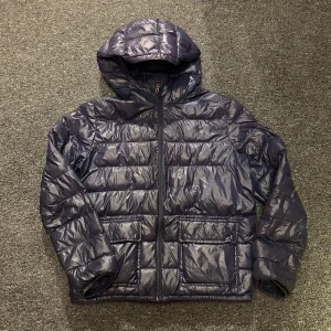 dunjacka från Polo Ralph Lauren - Snygg mörkblå dunjacka från Polo Ralph Lauren. Jackan har huva, två stora fickor framtill och dragkedja hela vägen upp. Klassisk quiltad look med broderad logga på bröstet. Jackan är i barnstorlek XL men sitter som S i vuxenstorlek