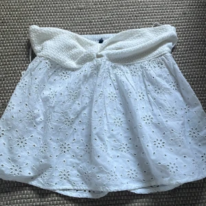 Vit broderad bandå topp - Supersöt vit kortkjol i bomull med broderade blommor och volang. Kjolen har en bred, smockad midja som ger en bekväm och snygg passform. Perfekt för varma dagar och ger en fräsch och somrig vibe.