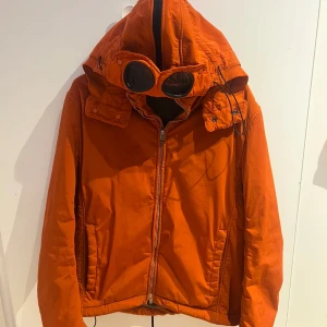 C.P. Company Jacka - Säljer en orange vindjacka från C.P. Company med ikoniska goggles i huvan. Jackan har dragkedja framtill, två sidofickor och justerbar huva. Materialet är lätt och passar perfekt för blåsiga dagar. Grym streetwear-vibe och riktigt snygg färg som sticker ut. OBS små deffekt som syns på bild 3