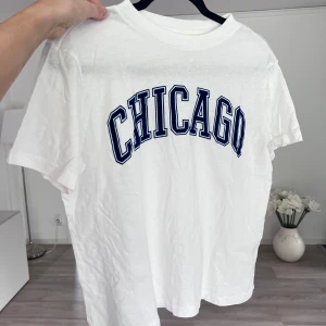 Vit t-shirt - Vit t-shirt från H&M med blå text 'CHICAGO' tryckt framtill i college-stil. Klassisk passform med rund hals och korta ärmar. Tillverkad i mjuk bomull, perfekt för en avslappnad och sportig look. Önskar du veta exakta mått? Tveka inte att höra av dig!