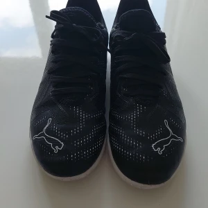 Svarta Puma fotbollsskor strl 35 - Snygga svarta fotbollsskor från Puma med vita detaljer och logga på både tå och häl. Skorna har snörning och perforerade partier för bättre ventilation. Yttermaterialet är syntet och sulan är platt och vit, perfekt för inomhusspel.