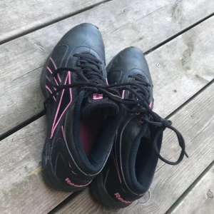 Svarta Reebok sneakers med rosa detaljer - Svarta Reebok sneakers med coola rosa detaljer och logga på sidan och hälen. Skorna har snörning och en unik sula med hexagonmönster i rosa och svart. Ovandelen är i syntetmaterial och textil, perfekt för dig som gillar sportig stil.