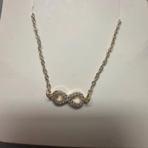Guldigt halsband med infinity-symbol - Snyggt halsband i guldton med en glittrig infinity-symbol dekorerad med små stenar. Kedjan är tunn och elegant, perfekt för att ge din outfit en extra touch. Låset är klassiskt och justerbart för bästa passform.