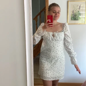 Vit blommig kortklänning med puffärm - Supersöt vit kortklänning med små blå blommor, långa puffärmar och söt knytning vid bysten. Klänningen har en feminin passform och är perfekt för varma dagar. Materialet känns lätt och luftigt, och den fyrkantiga urringningen ger en trendig vibe. Köparen står för frakt 📦 