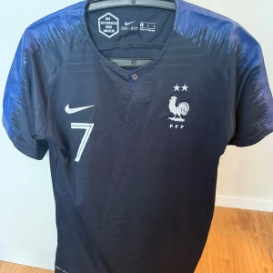 Frankrike Griezmann #7 Nike fotbollströja - Snygg officiell Frankrike landslagströja från Nike med Griezmann och nummer 7 på ryggen. Tröjan är mörkblå med blå detaljer på axlarna, har korta ärmar och är tillverkad i Dri-Fit material som andas. Perfekt för dig som älskar fotboll och vill bära ditt lag med stil.