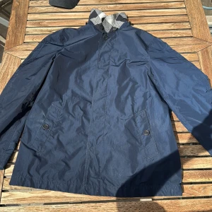 Marinblå burberry rock - Snygg marinblå vindjacka med klassiskt rutigt foder i kragen. Jackan har två fickor framtill med knappstängning och hög krage med knappar. Perfekt för blåsiga dagar och enkel att matcha med olika outfits.
