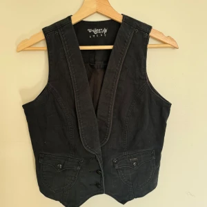 Svart jeansväst från College Clan - Svart jeansväst från med djup v-ringning och tre knappar framtill. Västen har två fickor med lock och knappdetaljer. Snygg, klassisk modell i denim som passar perfekt till lager-på-lager-stil.