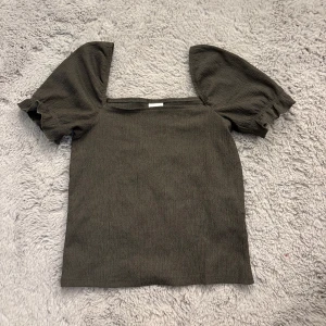 Mörkgrön topp med puffärmar - Supersnygg mörkgrön topp med fyrkantig urringning och puffiga korta ärmar. Tyget är lätt strukturerat och ger en cool känsla. Perfekt att styla med jeans eller kjol för en trendig look.