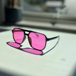 Svarta solglasögon med rosa glas - Coola solglasögon med svart båge och fyrkantiga rosa glas. Bågarna är i plast och har en klassisk pilotinspirerad form som ger en retrovibe. Perfekta för att sticka ut och ge din stil en färgklick.