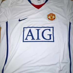  Vintage Manchester United vit bortatröja Nike - Vit Manchester United bortatröja från Nike med blå detaljer och klubbmärke på bröstet. Klassisk V-ringning med röd insida, AIG som sponsortryck på magen och blå Nike-logga. Tillverkad i lätt och ventilerande material, perfekt för fotboll eller samling.