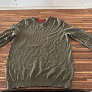 Olivgrön stickad tröja från Hugo - Snygg olivgrön stickad tröja från Hugo med rund halsringning och diskret broderad logga nertill. Tröjan har lång ärm och är i ett mjukt, tunt material som passar perfekt till lager-på-lager. Enkel och stilren design som funkar till många outfits. Köpt på room 1006 på Emporia för 2000kr. Den är liten i storleken så den passar även S.