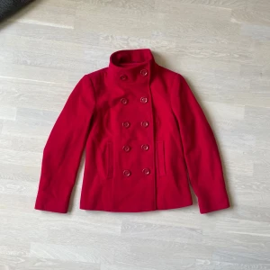 Röd skepparkavaj från Marks & Spencer - Snygg röd skepparkavaj från Marks & Spencer Portfolio med dubbelknäppning och hög krage. Jackan har långa ärmar, två fickor framtill och är tillverkad i ett mjukt tyg som känns lyxigt. Perfekt för dig som vill sticka ut med färg i höst och vinter.