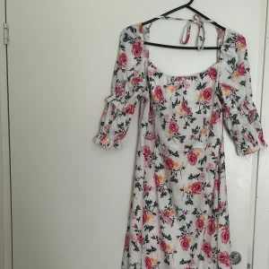 Vit långklänning med blommigt mönster - Superfin långklänning i vitt med rosa och gula blommor. Klänningen har trekvartsärmar med volangdetaljer och en söt knytning i nacken. Materialet känns lätt och flowy, perfekt för varma dagar. Den har en romantisk vibe och passar dig som gillar en feminin stil.
