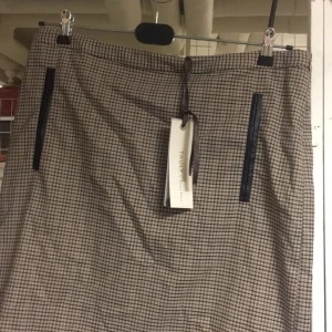 Rutig kjol från Part Two, stl 42 - Snygg rutig kjol från Truly by Part Two i beige och svart. Kjolen har raka linjer, två svarta detaljerade fickor framtill och klassiskt rutmönster. Perfekt för dig som gillar en stilren och tidlös look.