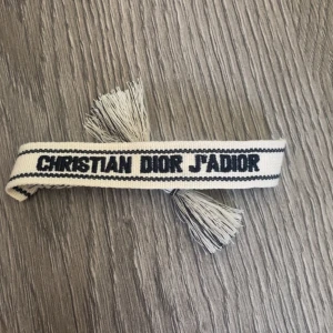 Christian Dior J'Adior armband - Snyggt armband från Christian Dior med texten 'J'Adior' broderad i svart på en vit bakgrund. Armbandet är gjort i vävd bomull och har coola tofsar i ändarna. 
