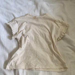 Beige topp med volangärm - Söt beige topp med korta ärmar och volangdetalj i ribbat material vid ärmsluten. Enkel och stilren design som passar perfekt till jeans eller kjol. Tillverkad i mjuk bomull för en skön känsla hela dagen.