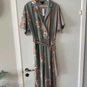 Ny Lindex grön jumpsuit, storlek M - 🌿Grön jumpsuit från Lindex med ett blommigt mönster i orange och rosa toner.  🌻Jumpsuiten har en skjortkrage, korta ärmar och ett knytband i midjan.  🌿Den har även fickor på sidorna och en dold knapp framtill. Perfekt för dig som gillar en avslappnad men snygg stil. 🚭Djurfritt och rökfritt hem.