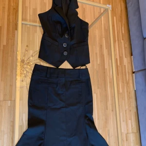 Svart kostymset med väst och kjol - Snyggt svart kostymset med ärmlös väst och figursydd kjol med volangkant. Västen har tre stora knappar och klassisk krage, samt fickdetaljer framtill. Kjolen har hällor för skärp och dekorativa fickor bak. Perfekt för dig som vill sticka ut med en modern twist. Xs/S