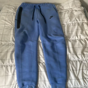 Blå mjukisbyxor från Nike, storlek M - Snygga ljusblå mjukisbyxor från Nike med svarta snören i midjan och en svart dragkedjeficka på ena benet. Byxorna har ribbade muddar vid bensluten och är tillverkade i mjukt bomullsmaterial. Perfekta för chill eller träning.