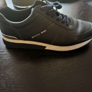Svarta sneakers från Michael Kors - Stilrena svarta sneakers från Michael Kors i skinn med vita detaljer på sulan och diskret logga i silver på sidan. Klassisk rund tå, snörning och bekväm platt sula. Perfekta för en trendig och sportig look.