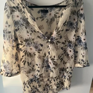 Blommig blus med knappar och v-ringning - Supersöt blommig blus från Vero Moda i ljusbeige med blommor. Den är genomskinlig  och V-ringad med små knappar framtill och smockad nederkant. Lätt och luftig med trekvartsärm, perfekt för vår och sommar. 
