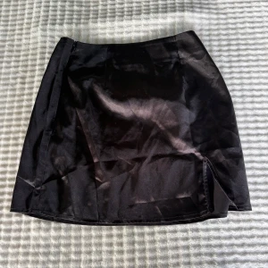 Svart kortkjol med slits från SHEIN - Snygg svart kortkjol från SHEIN i glansigt material. Kjolen har en enkel design med en liten slits framtill som ger en trendig touch. Perfekt att matcha med en crop top eller oversized tröja för en cool look.