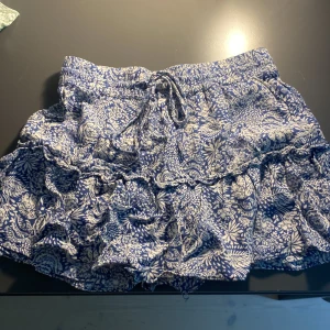 Blåvit volangkjol med snörning - Supersöt kort kjol i blått och vitt med blommigt mönster och volanger. Det haf inbyggda shorts.
