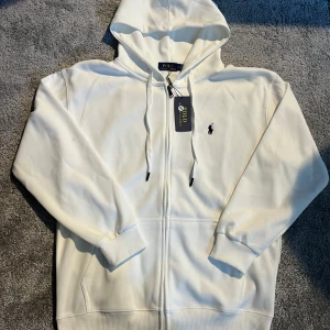 Vit hoodie från Polo Ralph Lauren - Snygg vit hoodie från Polo Ralph Lauren med dragkedja och klassisk broderad logga på bröstet. Tröjan har huva med snörning, kängurufickor och ribbade muddar. Perfekt för en clean och stilren look. Materialet är mjuk bomull som känns skönt mot huden.