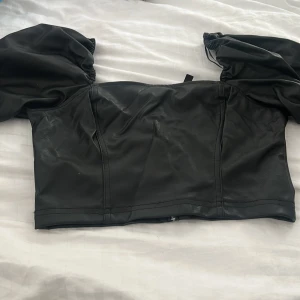 Svart croppad skinnkorsett - Säljer en svart croppad korsettopp i skinnimitation med puffiga korta ärmar. Toppen har en snygg figurnära passform och markerade sömmar framtill som ger en edgy vibe.