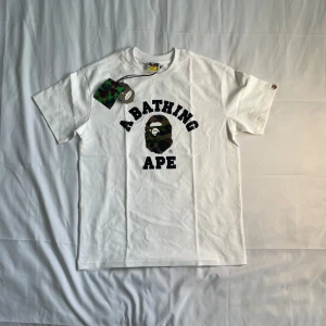 Vit Bape T-shirt - A Bathing Ape (BAPE) College-T-shirt – Vit Camo-huvudlogotyp (Storlek M)  Helt ny, oanvänd, med originaletiketter. Ikonisk BAPE-t-shirt med college-logotyp och det signaturkamouflage-aphuvudet på skarp vit bomull.  • Storlek: Medium • Skick: Nytt med etiketter • Färg: Vit med kamouflage-/logotyptryck • Passform: Normal BAPE-passform  🔥 Ett måste för alla streetwear-kollektioner eller BAPE-fans.  Skickas snabbt – skicka ett meddelande om du har några frågor!