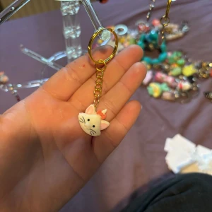 Nyckelring med söt katt i vitt och rosa - Supergullig nyckelring med en vit katt med rosa rosett och svarta detaljer. Katten har en rund form och hänger i en guldfärgad kedja och ring. Perfekt accessoar till väskan eller nycklarna för dig som älskar söta djurmotiv.