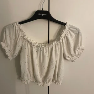 Vit broderad blus från Gina Tricot - Supersöt vit blus från Gina Tricot med broderade detaljer och små hålmönster. Modellen är croppad med resår i midja, ärmslut och halsringning. Puffiga korta ärmar och offshoulder-stil ger en romantisk vibe. Perfekt för varma dagar och sommarhäng!