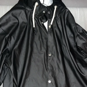 Svart regnjacka med huva och snörning - Svart regnjacka med huva och vita snören, perfekt för blöta dagar. Jackan har tryckknappar framtill och en stilren look som funkar till det mesta. Materialet är syntet och jackan har en loose passform för extra komfort. Som nytt 